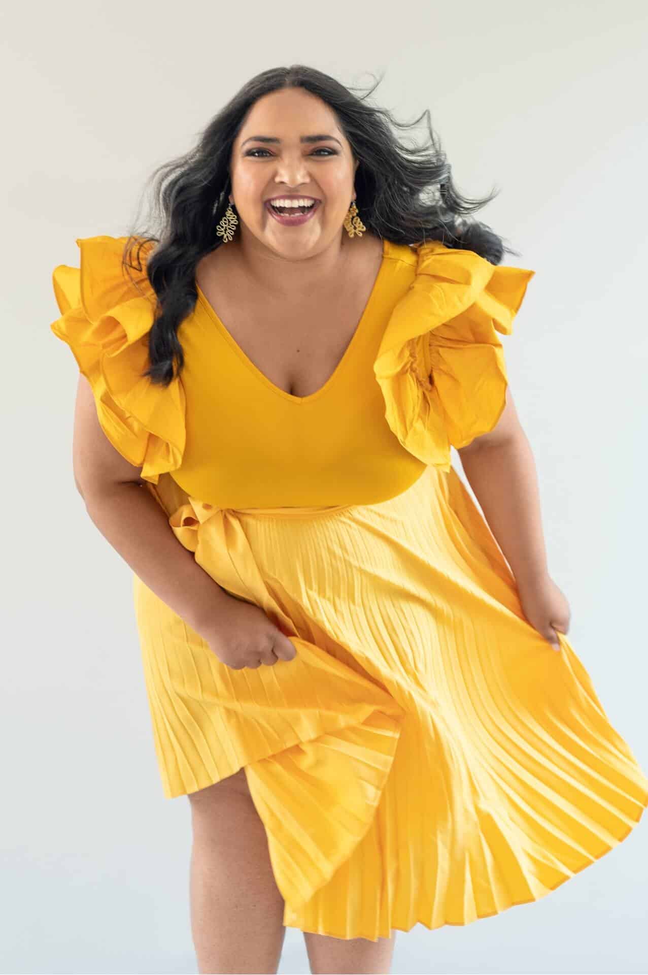 In studio portrait. The image features a woman in a bright yellow dress looking at the camera as she dances and sways her skirt.