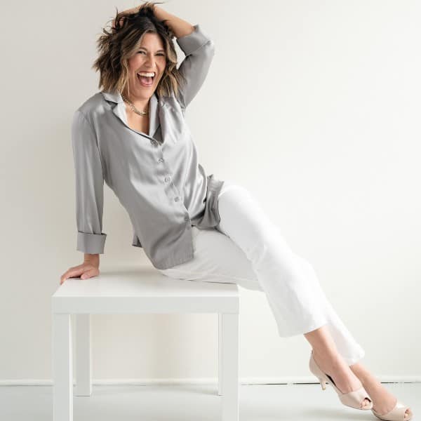 In studio 40OVER40 portrait. Woman in a silver silk top and white pants is seated on a white table, posed against a white background. She is ruffling her hair and looks jovial.